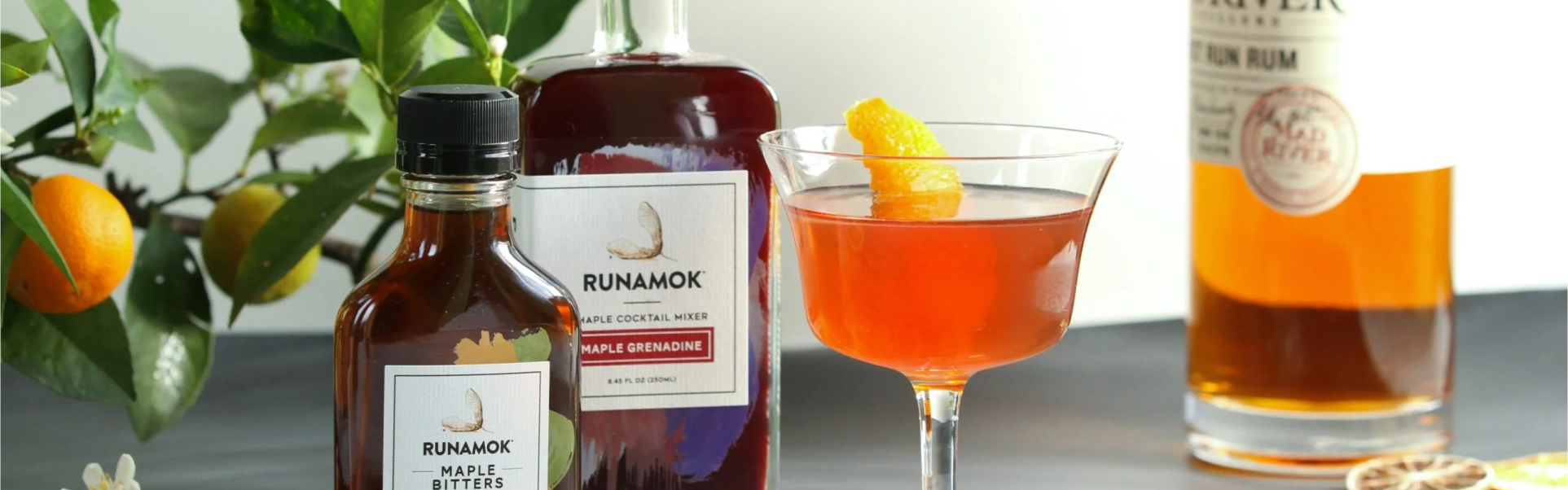 2 different bottles of Runamok products and a cocktail glass, lemons and a Rum bottle on the background